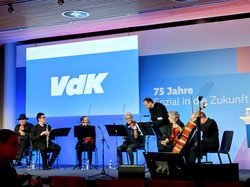 Orchestermusikerinnen und Musiker auf der Bühne vor blauer Leinwand mit der Aufschrift VdK 75 Jahre "Sozial in die Zukunft". Klick öffnet eine vergrößerte Ansicht.